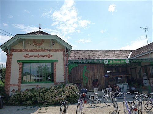 Een charmant gebouw met gedetailleerd metselwerk naast een houten schuur met een fietslogo in Arçais, Frankrijk. Fietsen zijn geparkeerd buiten in de zon