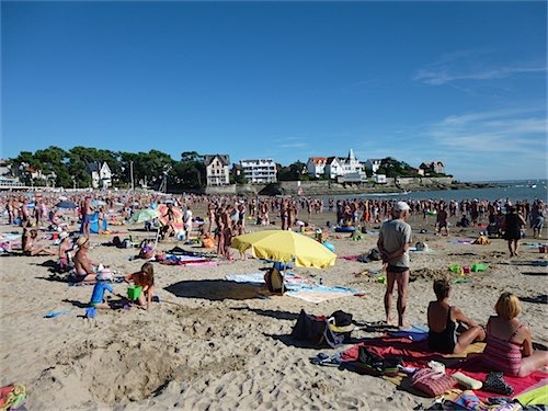 Drukte op het zonnige strand nabij de haven van Royan, Frankrijk. Vakantiegangers genieten van de zon en zee, terwijl op de achtergrond karakteristieke gebouwen de kustlijn sieren.