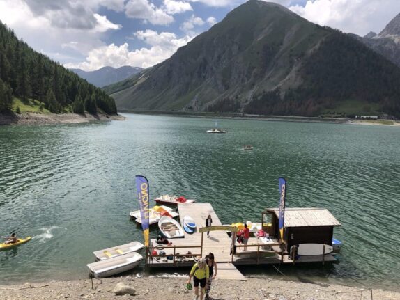 Een schilderachtig berglandschap met het Lago di Livigno in het midden. Watersportactiviteiten en kajakken zijn zichtbaar op de voorgrond met een houten steiger, kajaks, boten en enkele mensen. Er zijn ook vlaggen met reclame te zien. In de verte zijn weelderige groene bossen en majestueuze bergpieken zichtbaar onder een lichte wolkendek.