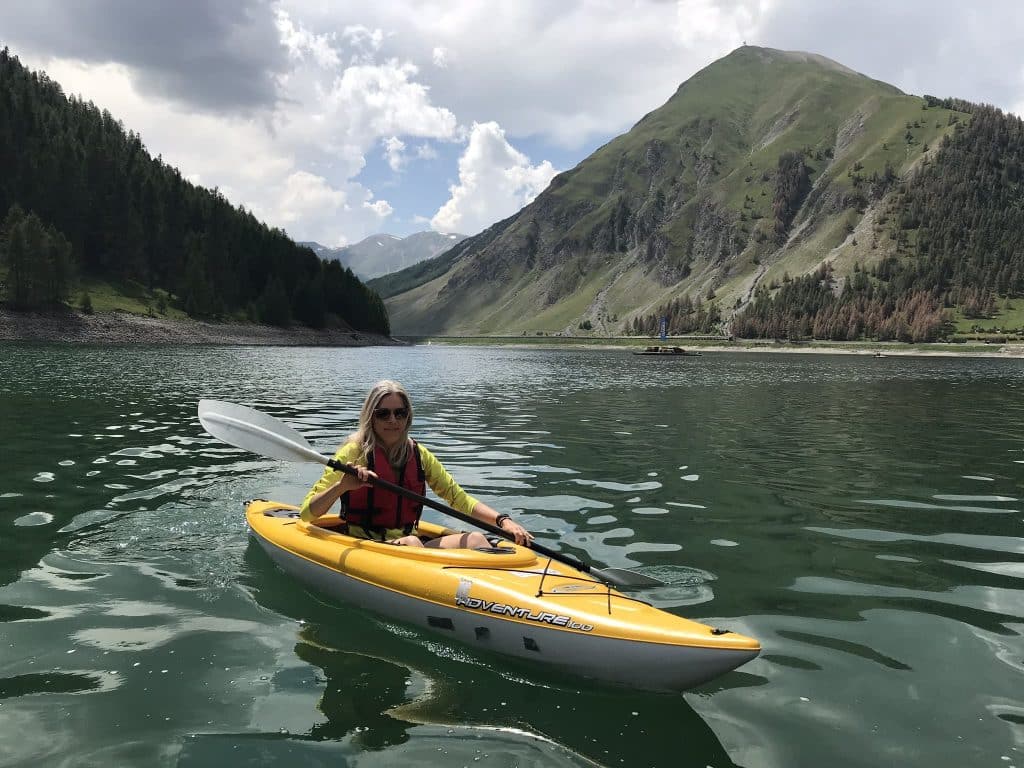 Een vrouw in een gele kajak op het Lago di Livigno, actief deelneemt aan watersportactiviteiten. De achtergrond toont weelderige groene bossen en indrukwekkende bergtoppen die het serene water omringen. De vrouw draagt een reddingsvest en hanteert een peddel terwijl ze op het kalme meer kajakt.