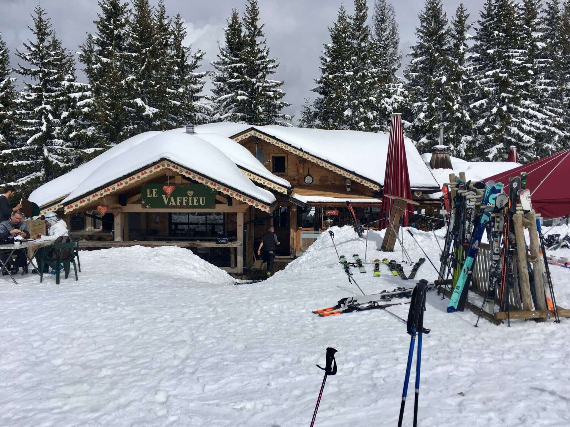 Een gezellige berghut genaamd 'Le Vaffieu' in het skigebied Portes du Soleil, gelegen te midden van besneeuwde bomen. Buiten de hut zijn skiërs te zien die genieten van hun maaltijd aan houten tafels. Diverse ski's en skistokken zijn geparkeerd in de sneeuw in de voorgrond.