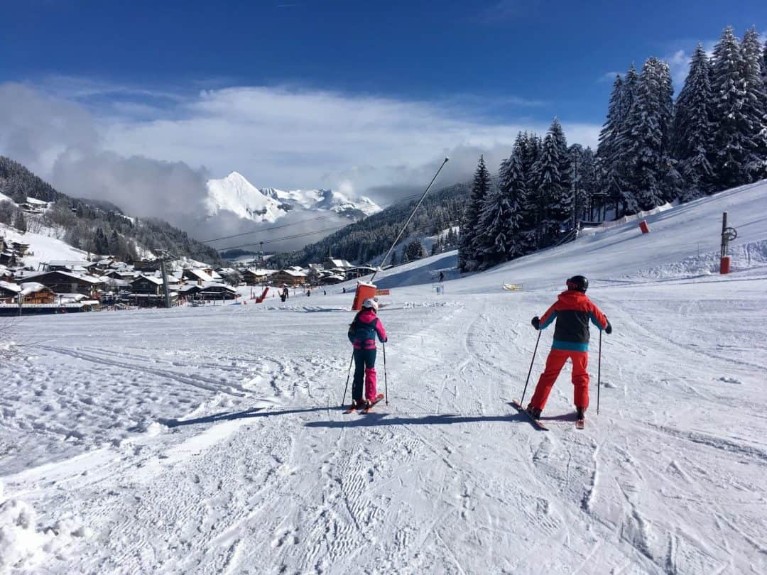 Twee skiërs, één in roze en de ander in oranje skikleding, staan klaar om een afdaling te maken op de skipiste. Op de achtergrond is een schilderachtig Alpendorp te zien, omgeven door besneeuwde bergen en met helderblauwe lucht erboven, in het skigebied Portes du Soleil