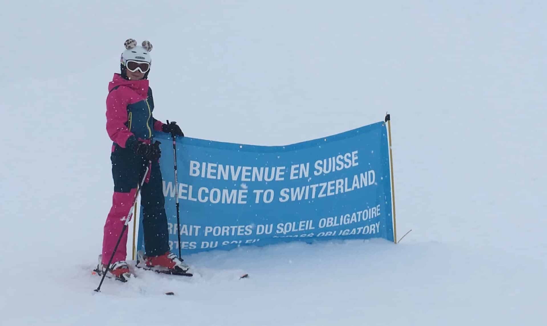 Een skiër in roze skikleding, voorzien van een skibril en helm met decoratieve oren, staat naast een blauwe banner met de tekst 'Bienvenue en Suisse - Welcome to Switzerland' in het skigebied Portes du Soleil te midden van een sneeuwstorm