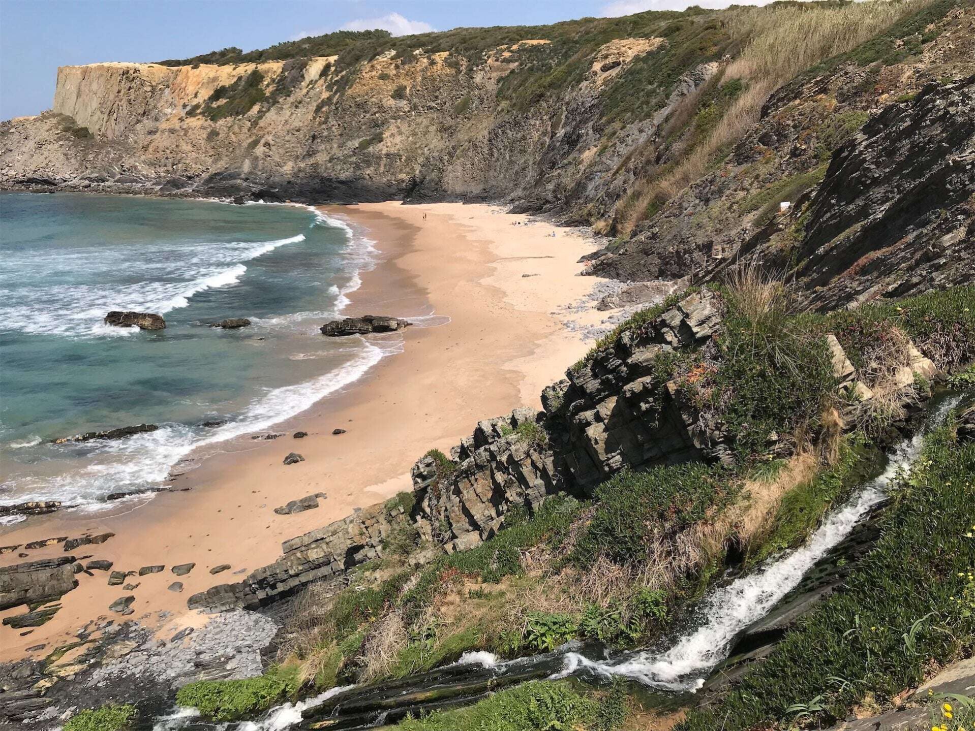 Een adembenemend uitzicht op een afgelegen strand in Portugal, omgeven door steile kliffen en groene vegetatie. Heldere turquoise golven spoelen aan op het gouden zand, terwijl een kleine waterval vanaf de klif naar beneden stroomt. Dit is een scène langs de wandelroute Rota Vicentina