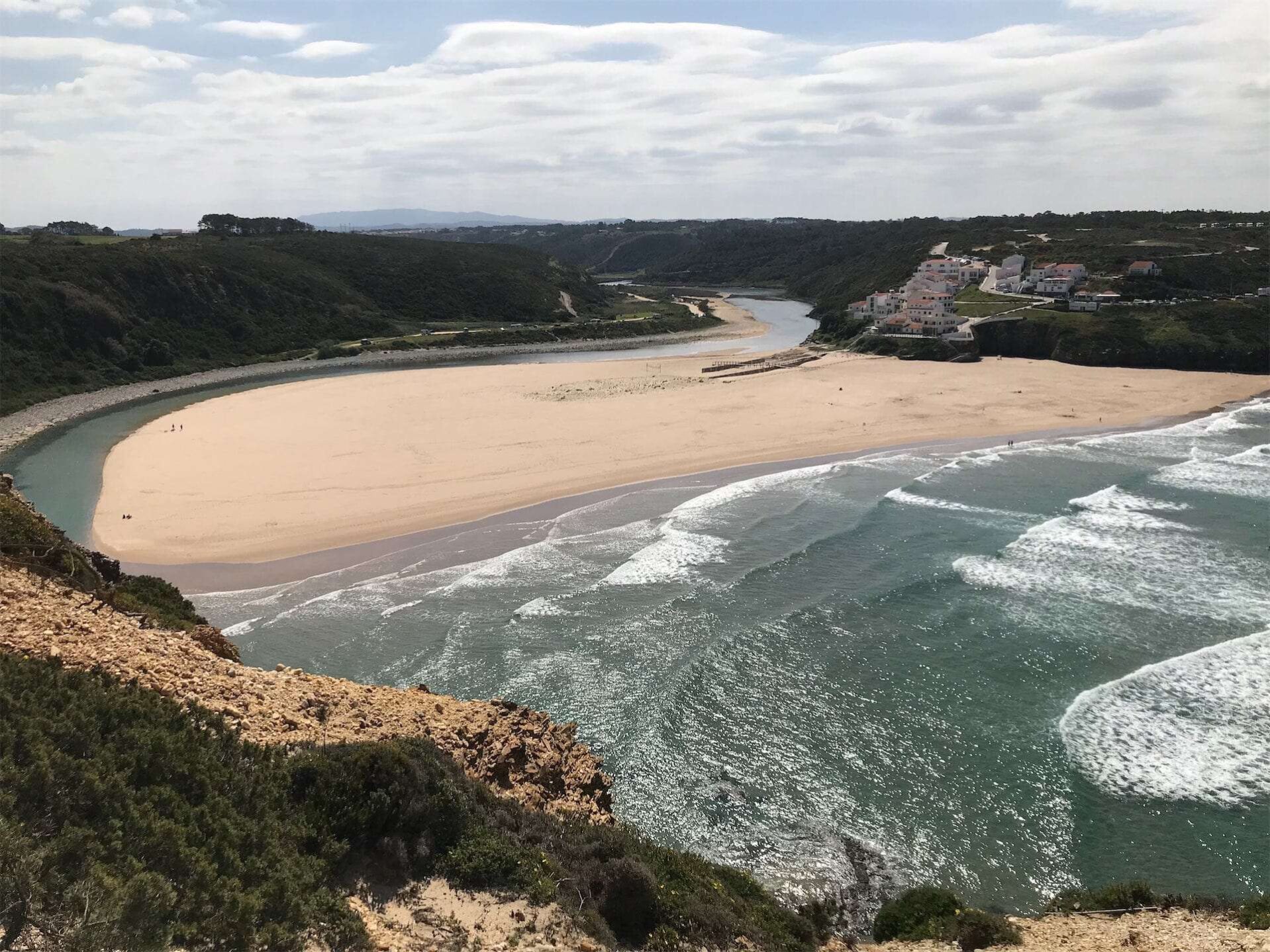 Panoramisch uitzicht op een serene baai aan de Rota Vicentina-route met een uitgestrekt zandstrand, schuimende golven en een cluster van gebouwen aan de kustlijn, omgeven door groene heuvels en kliffen.