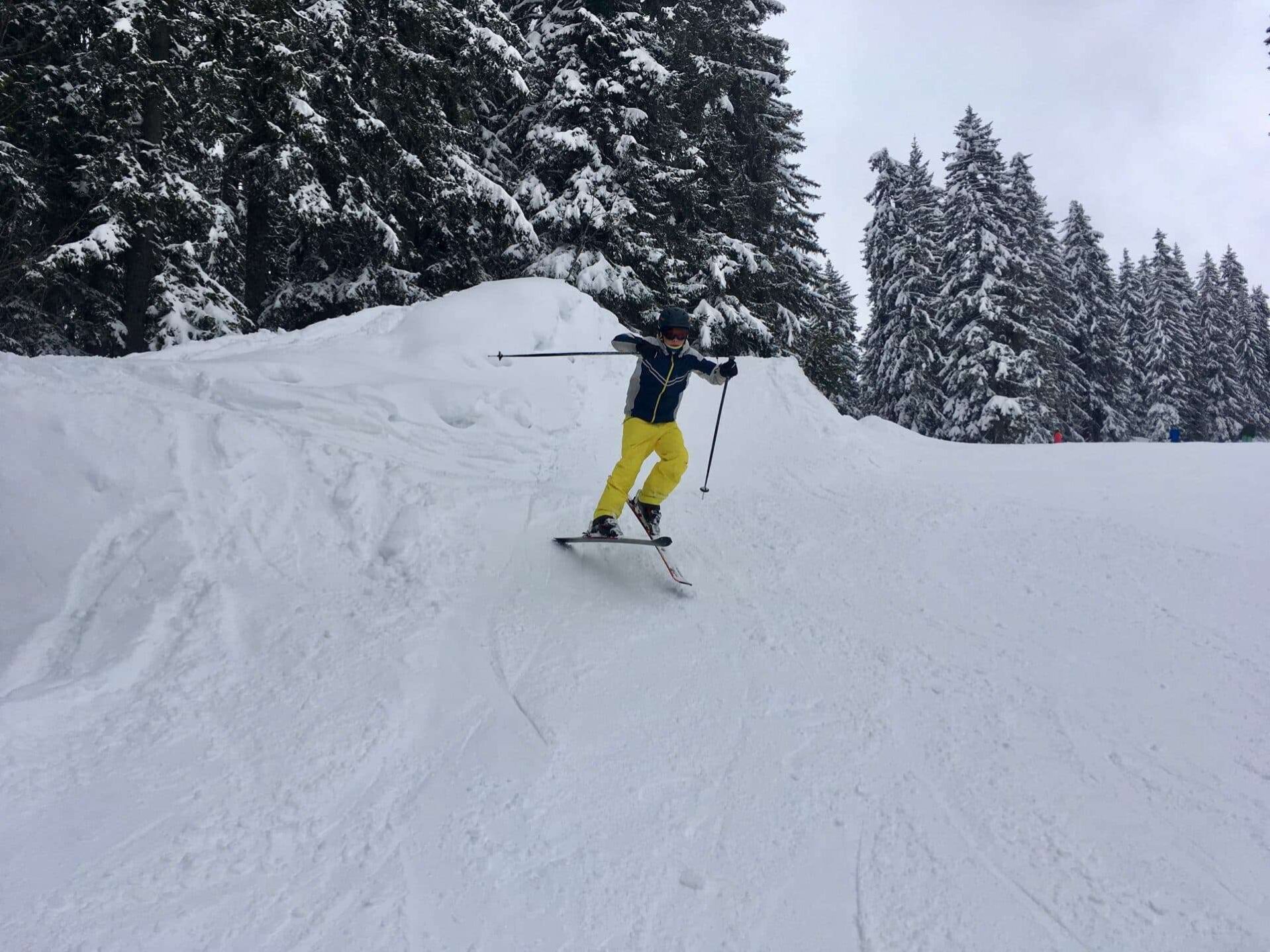 Een skiër in een geel en blauw skipak maakt een bocht op een besneeuwde helling in het skigebied Portes du Soleil, omgeven door dichtbeboste, met sneeuw bedekte dennenbomen. In de verte zijn andere skiërs zichtbaar tussen de bomen.