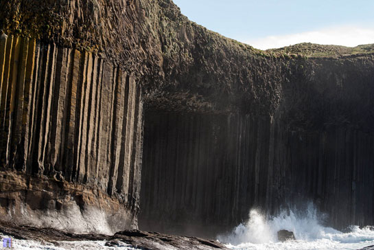 De foto toont de indrukwekkende basaltzuilformaties van Staffa, een eiland in Schotland. Krachtige golven beuken tegen de rotsachtige kust, waarbij opspattend water zichtbaar is. De verticale kolommen van de kliffen steken opvallend af tegen de achtergrond van de heldere lucht en het omringende landschap.