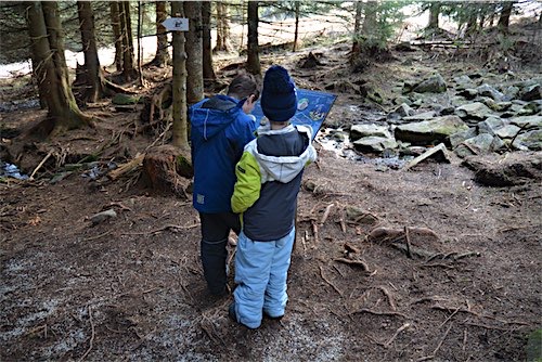 Twee jonge jongens in warme winterkleding staan in een bos in de Vogezen, Frankrijk, met op de grond gevallen bladeren en stenen. Ze bekijken samen een kaart die een van hen vasthoudt. Ze staan bij een paal met een bordje erop, mogelijk een wegwijzer of informatiebord.