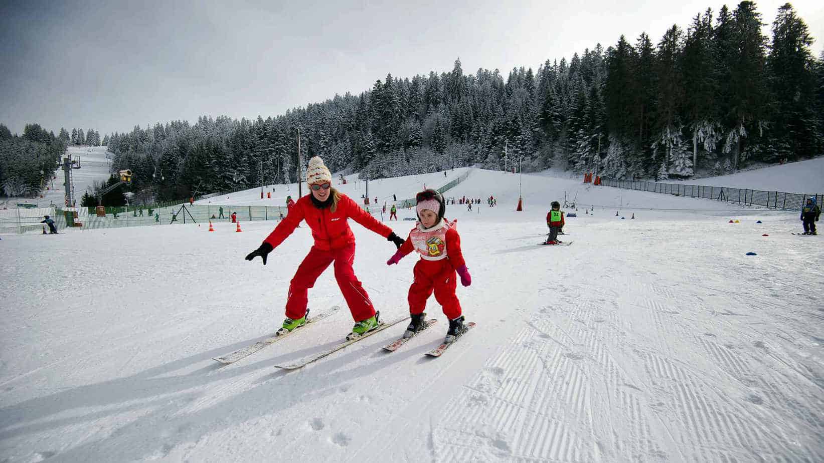 Een volwassene en een kind, beiden gekleed in rode skikleding, staan op ski's en lachen op de besneeuwde hellingen van de Vogezen in Frankrijk. De sfeer is levendig met andere skiërs op de achtergrond, skiliften en met sneeuw bedekte bomen.
