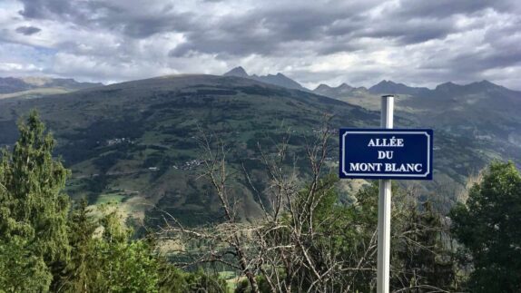Een uitkijkpunt met uitzicht op de glooiende groene heuvels en verre pieken van de Franse Alpen, onder een bewolkte hemel. Op de voorgrond staat een blauw bord met de tekst 'ALLÉE DU MONT BLANC', bevestigd aan een paal.