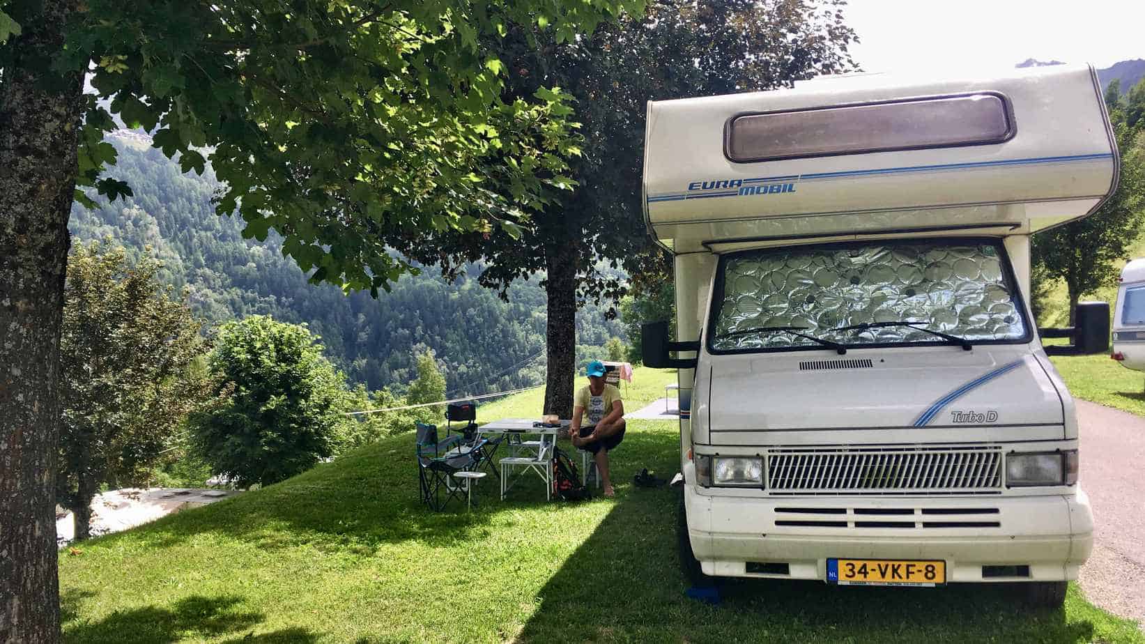 Een camper geparkeerd onder een schaduwrijke boom met een persoon ernaast, omgeven door het groene landschap van de Franse Alpen.