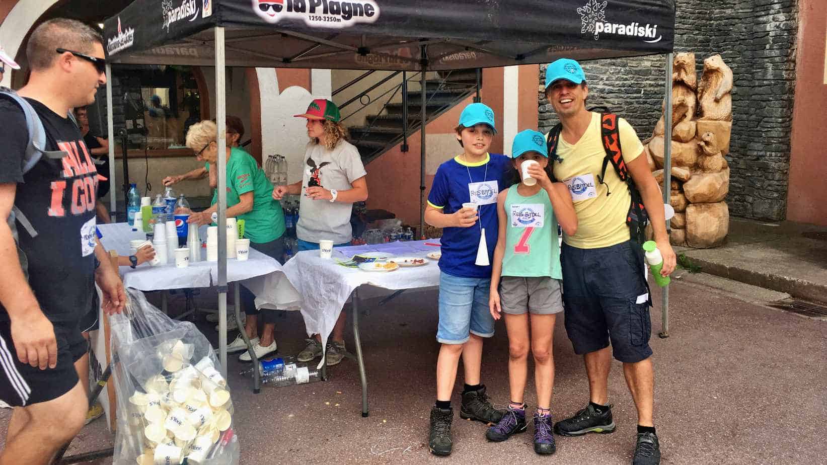 Een groep mensen, waaronder kinderen in matchende petten, staan bij een stand met het opschrift 'La Plagne' en 'Paradiski', waar een dame drankjes serveert.