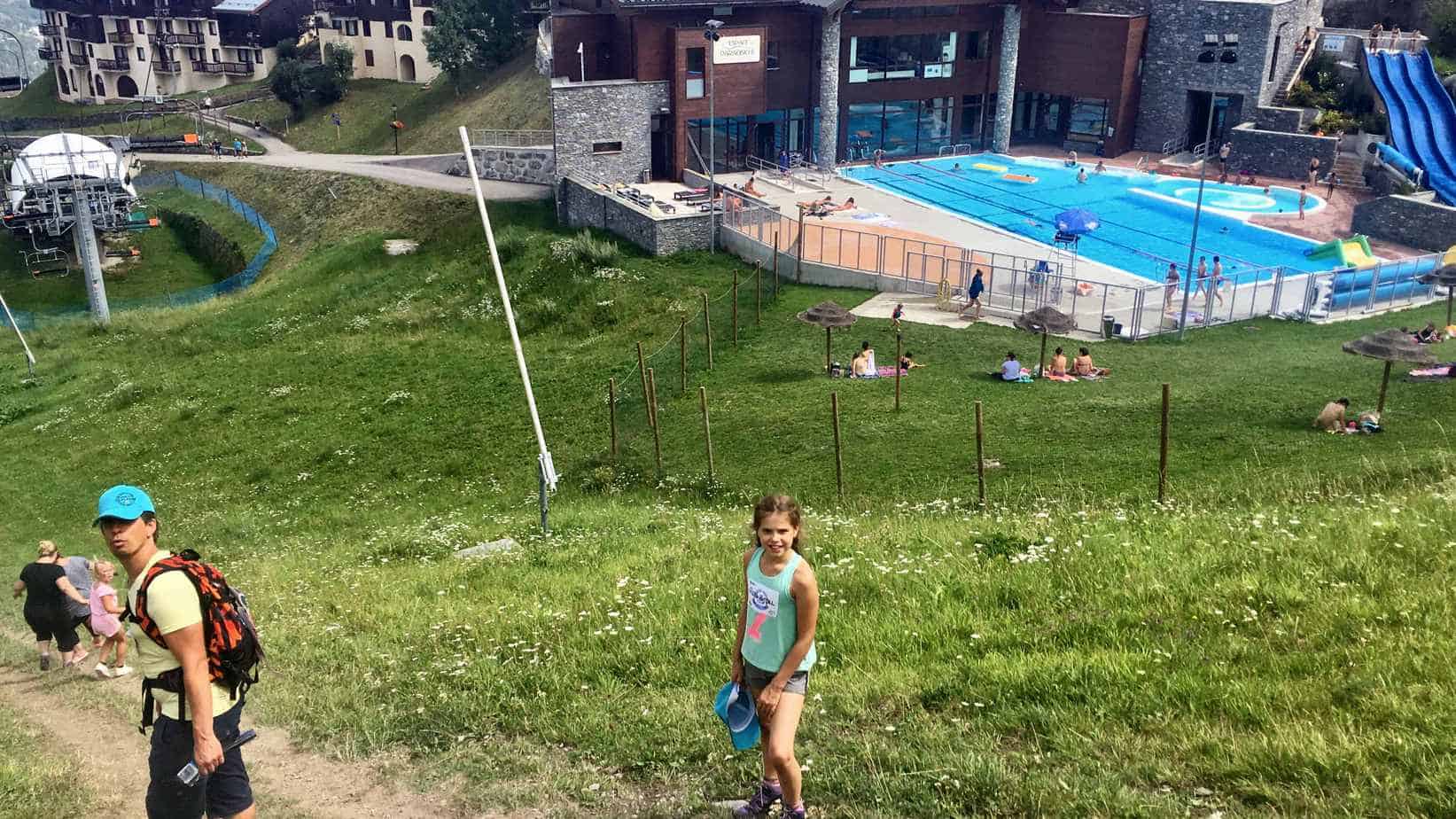 Een openluchtzwembad in La Plagne, Frankrijk, omgeven door groen gras met mensen die zonnen en zwemmen. Voorgrond toont een jongen met een pet en rugzak en een meisje in een groen hemd, beiden wandelend op een pad. Op de achtergrond zijn gebouwen en een stoeltjeslift zichtbaar.