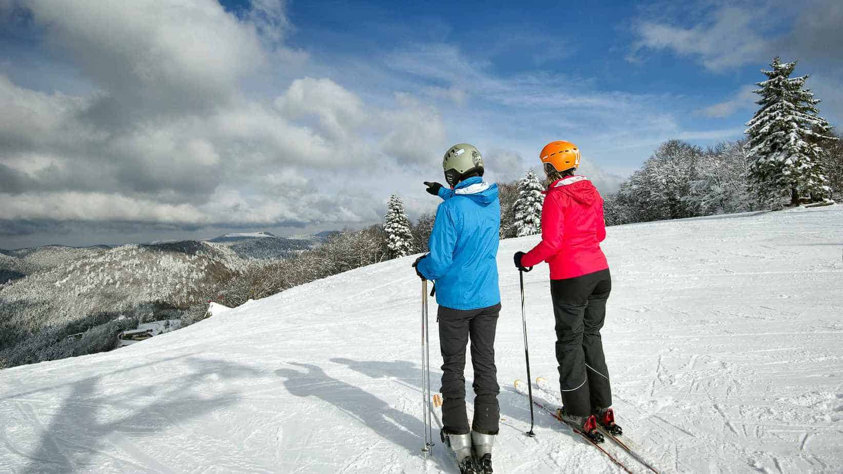 Twee skiërs, één in een blauwe jas en de andere in een roze jas, staan bovenaan een besneeuwde helling. Ze kijken uit over een indrukwekkend berglandschap bedekt met sneeuw. De lucht is gedeeltelijk bewolkt en er zijn sporen van andere skiërs zichtbaar in de sneeuw voor hen.
