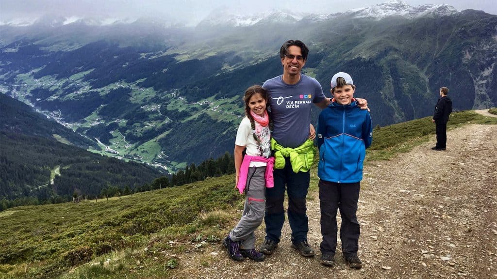 Een glimlachende man met een zonnebril en een grijs T-shirt staat op een bergpad in Ischgl, Oostenrijk, met aan weerszijden twee opgetogen kinderen. Het meisje draagt een witte top met een felroze vest, terwijl de jongen een blauwe jas draagt. Op de achtergrond ontvouwt zich een adembenemend uitzicht op de groene alpenvallei, doorsneden door een slingerende rivier. Rechts staat nog een persoon, blik gericht op de majestueuze bergen die gehuld zijn in wolken, wat de foto een gevoel van avontuur en familieband geeft tijdens een actieve zomervakantie.