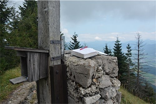 Gipfelbuch ligt op een stenen sokkel bij een houten paal op de top van een berg met uitzicht op een bosrijke helling in de mist."