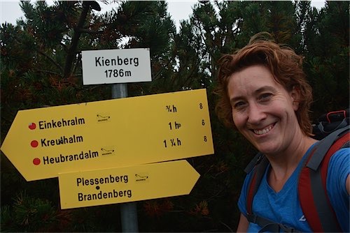 Een blije wandelaar met een rode rugzak naast een gele wegwijzer op de Kienberg op 1786 meter hoogte, met richtingen naar verschillende bestemmingen.