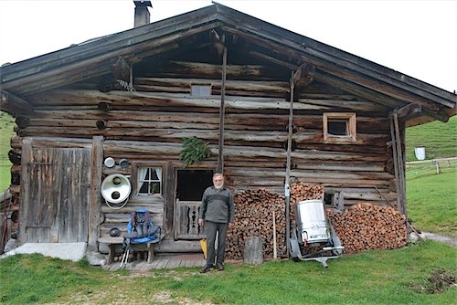 Persoon staat naast een rustieke houten berghut met een opgestapelde houtvoorraad en huiselijke voorwerpen aan de buitenmuur.