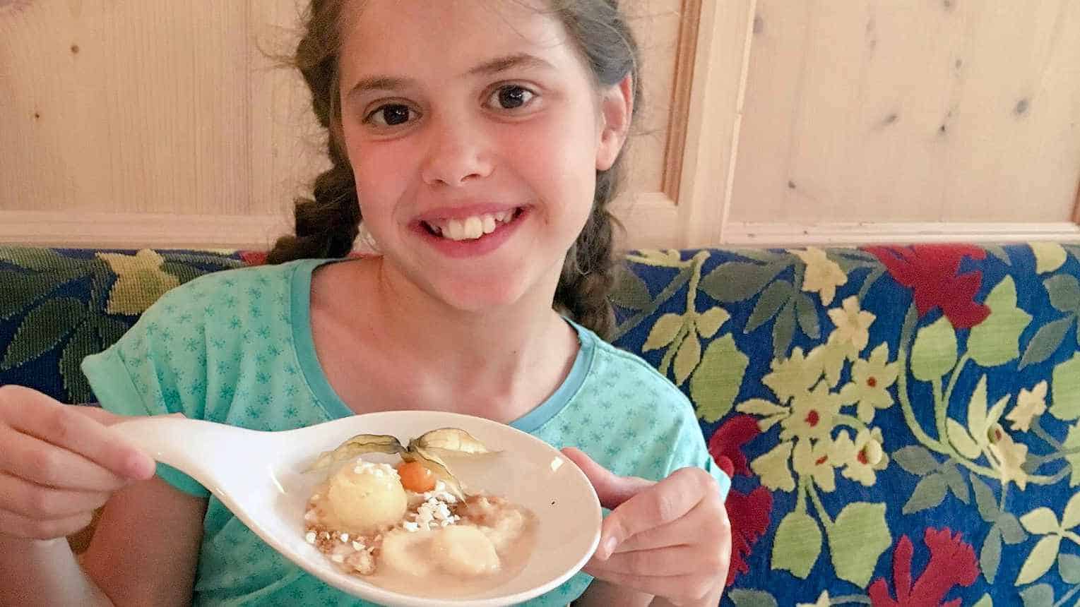 Een vrolijk jong meisje presenteert trots een lepel met een verleidelijk dessert in Hotel Ischglerhof, Ischgl. De lepel bevat een bolletje ijs en wat vruchten, een zoete afsluiter na een dag vol avontuur in de Oostenrijkse Alpen.