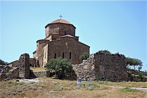 Een indrukwekkende oude stenen kerk gelegen op een heuvel in Georgië, omringd door ruïnes en muren. Enkele bezoekers bewonderen het bouwwerk van dichtbij, terwijl de heldere blauwe lucht het tafereel complementeert