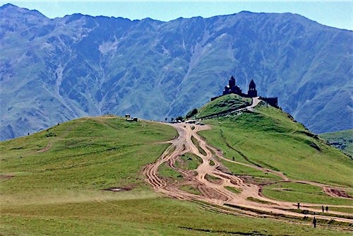 Een schilderachtig zicht op een klooster dat trots bovenop een groene heuvel in Georgië staat, met kronkelende paden die naar de top leiden. De majestueuze bergketens op de achtergrond versterken de serene sfeer van deze bijzondere plek, populair bij reizigers van Better Places.