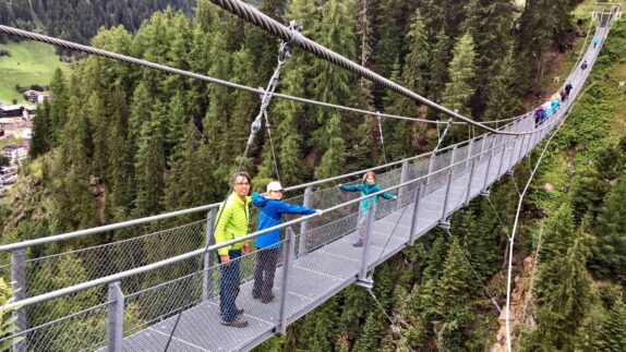 Drie personen, vermoedelijk een familie, staan op een hangbrug omgeven door weelderige groene dennenbossen in Ischgl, Oostenrijk, genietend van een actieve zomervakantie en de majestueuze natuur.