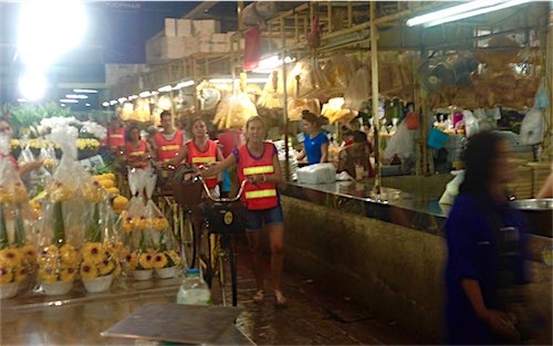 Een groep fietsers in opvallende vesten loopt naast hun fietsen in een drukke nachtmarkt in Bangkok, vol met bloemenstalletjes en verkopers, verlicht door bovenliggende lampen.