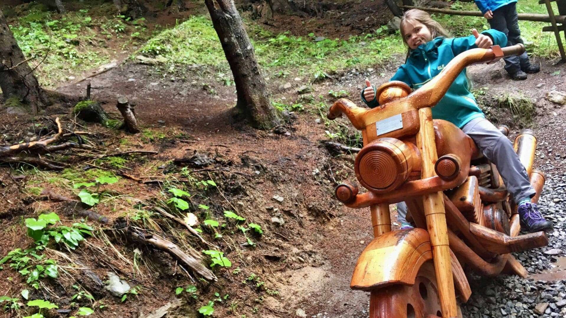 Een kind, met een grote glimlach en een duim omhoog, geniet van een creatieve houten speelstructuur in de vorm van een motorfiets langs een kindvriendelijke wandelroute in Ischgl, Oostenrijk. De route, omgeven door een bosrijke omgeving met rijke vegetatie en een vochtige ondergrond, nodigt uit tot ontdekken en spelen, wat de natuurlijke nieuwsgierigheid van kinderen stimuleert.