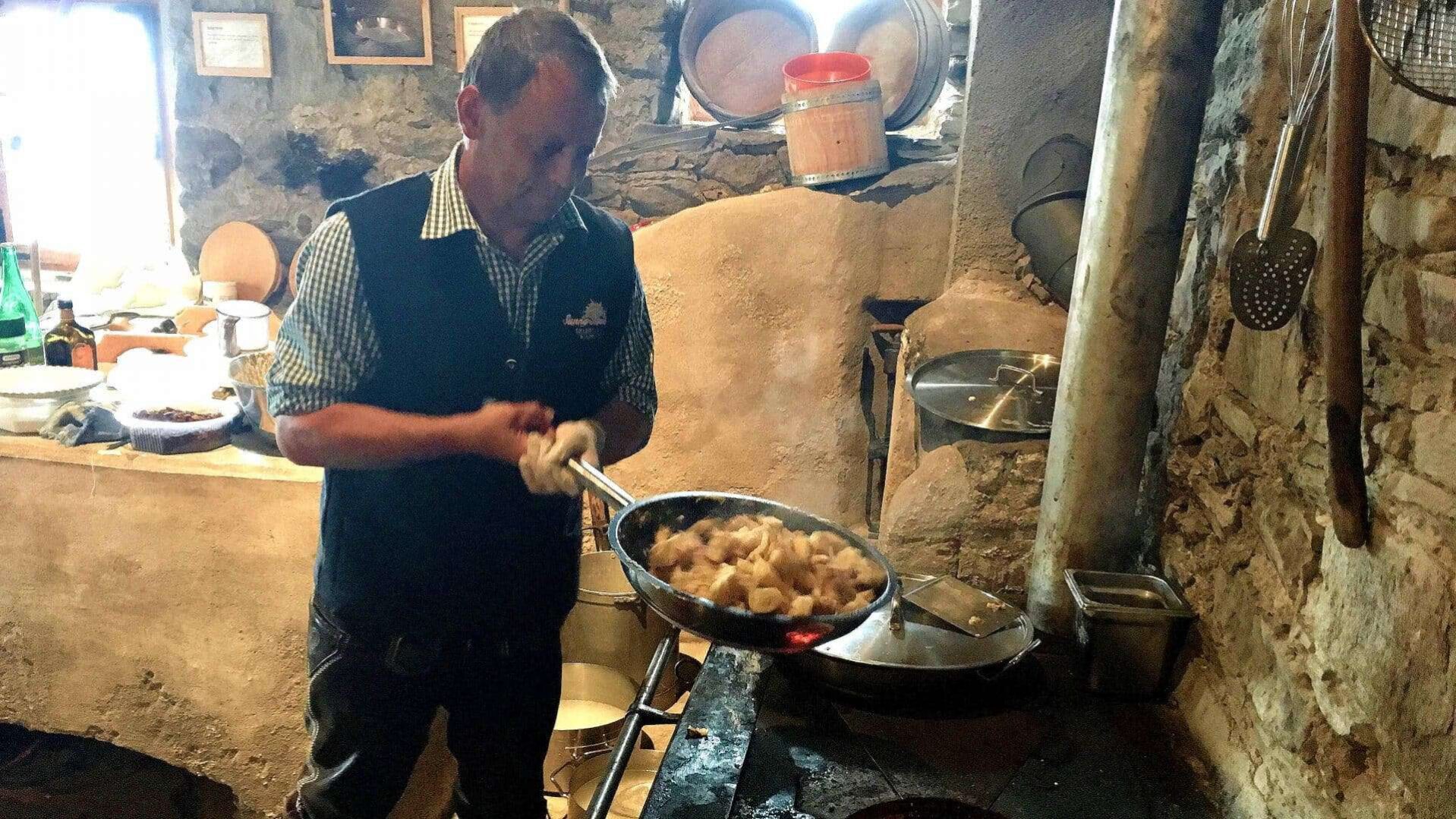 Een man, gekleed in een traditioneel gilet met een geruit overhemd, is geconcentreerd aan het koken in een authentieke, rustieke keuken in Ischgl, Oostenrijk. Hij hanteert een grote koekenpan boven een open vuur, waarin stukjes vlees sudderen en stomen. De keuken is ingericht met stenen muren en houten keukengerei hangt binnen handbereik, wat bijdraagt aan de sfeer van een traditionele Oostenrijkse kookworkshop.