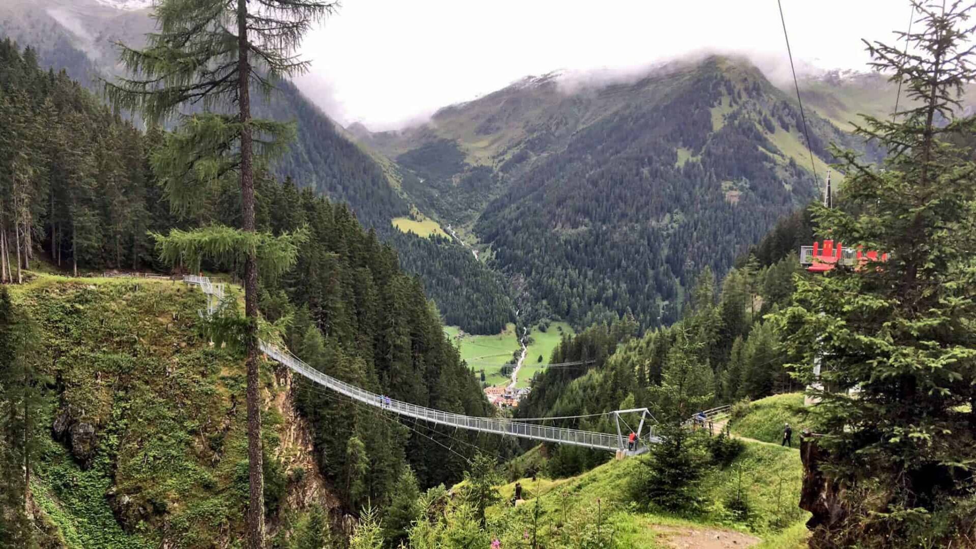 Drie wandelaars, twee volwassenen en een kind, genieten van het uitzicht terwijl ze op een indrukwekkende hangbrug in Ischgl, Oostenrijk staan. De brug, die hoog boven een groene kloof uitstrekt, biedt een avontuurlijke route tussen de dichte naaldbossen, en geeft een panoramisch uitzicht op het dorpje beneden.