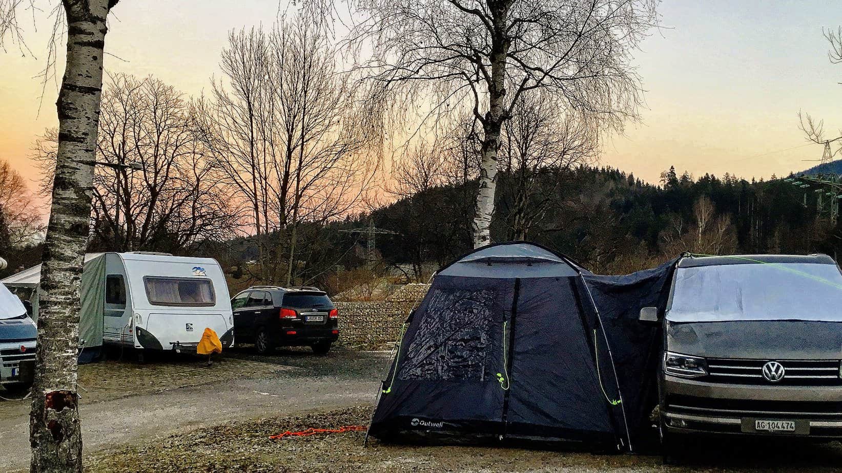 Een kampeerterrein in Oostenrijk tijdens de schemering met een roze-gouden lucht op de achtergrond. Campers, auto's en een tent zijn te zien, omringd door kale bomen en een berkenboom op de voorgrond. Een Volkswagen busje is geparkeerd naast een tent, beide bedekt met een dunne laag vorst.