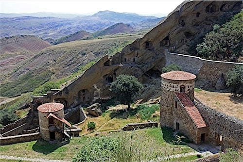 Een oude Georgische nederzetting met stenen gebouwen en torens op een heuvelachtig terrein, met op de achtergrond de glooiende heuvels van Georgië. De wegen slingeren door het landschap, waar Marshrutka's vaak reizen.