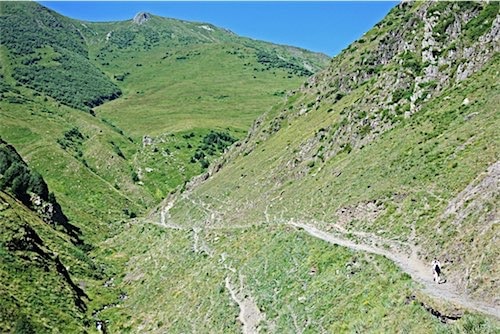 Een schilderachtig uitzicht op een kronkelend wandelpad in het groene berglandschap van Georgië. Op het pad is een eenzame wandelaar zichtbaar en in de verte kunnen we de kenmerkende Marshrutka's van Georgië herkennen.