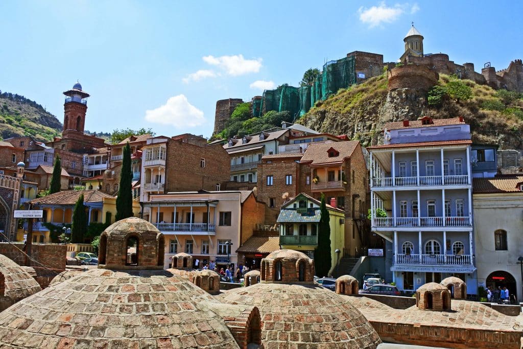 Een levendig straatbeeld in Tbilisi, Georgië, waar traditionele gebouwen zich vermengen met oude koepelvormige structuren. In de verte verheffen historische torens en stadsmuren zich op een heuvel, waardoor het rijke erfgoed van de stad wordt benadrukt.