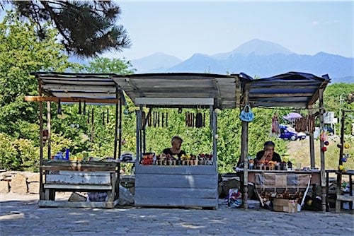 Twee lokale verkopers in Georgië zitten in hun kraampjes onder schaduwrijke afdakjes, waar ze diverse producten uitstallen. Op de achtergrond zijn de majestueuze bergen van Georgië zichtbaar en een pad waar Marshrutka's frequent langsrijden.