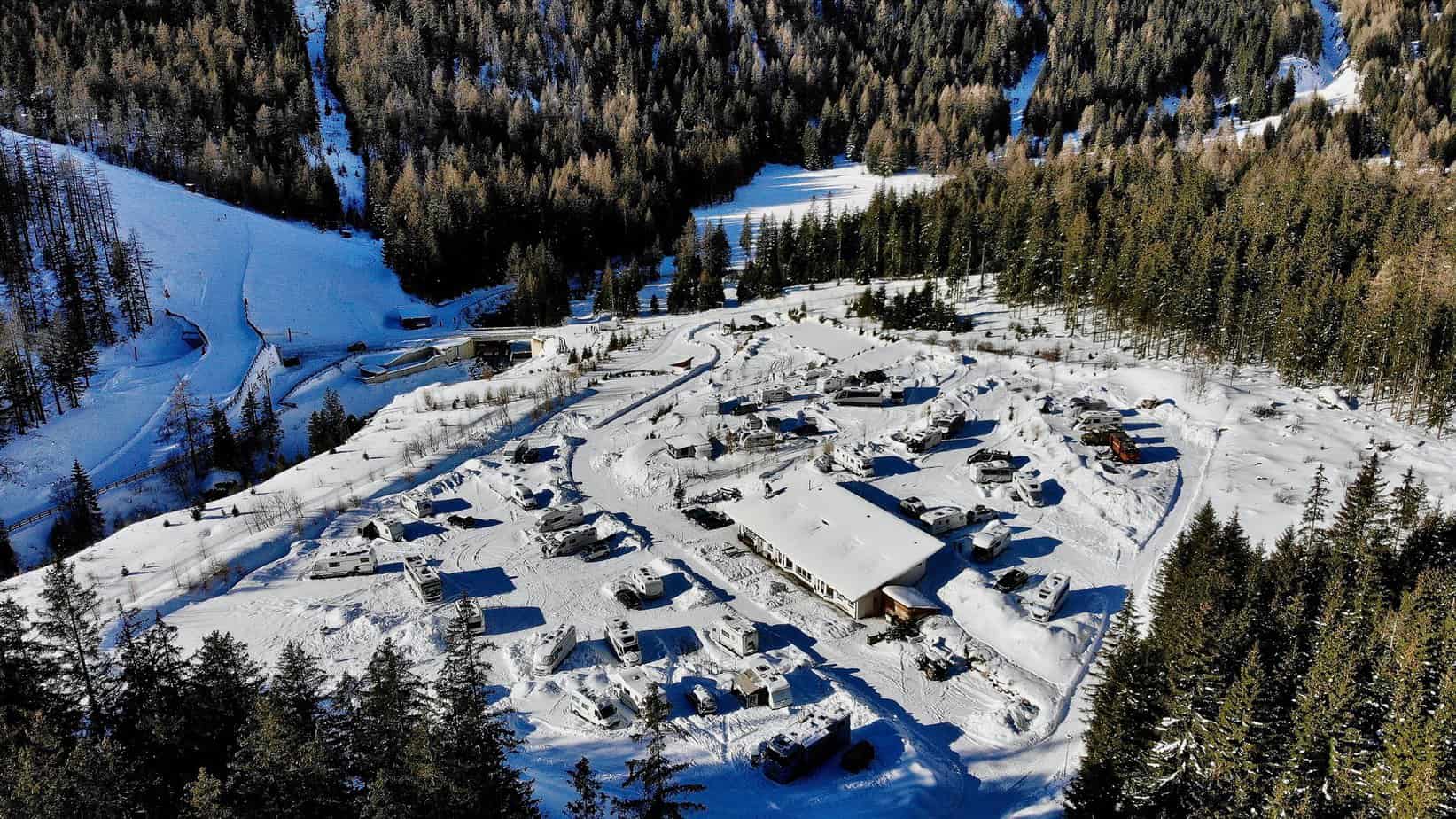 Een luchtfoto van een besneeuwd camperterrein genesteld tussen de met dennenbomen bedekte heuvels in Oostenrijk. Verschillende campers zijn geparkeerd op het terrein, omringd door een dik tapijt van sneeuw, terwijl de winterzon de bomen en het landschap verlicht.